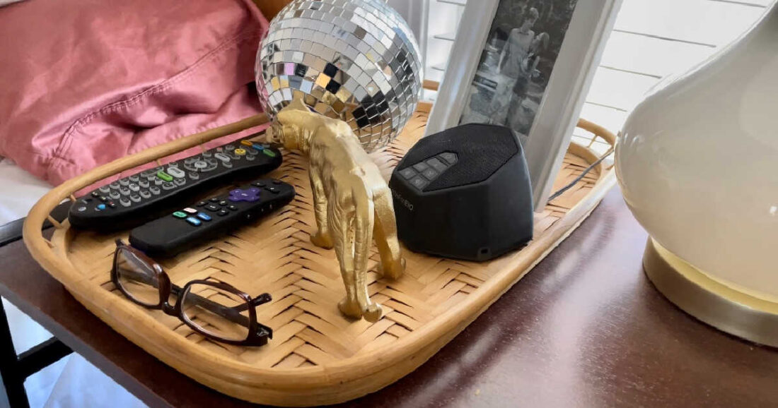 Nightstand with rattan tray organized with remotes, glasses, photo and decor.